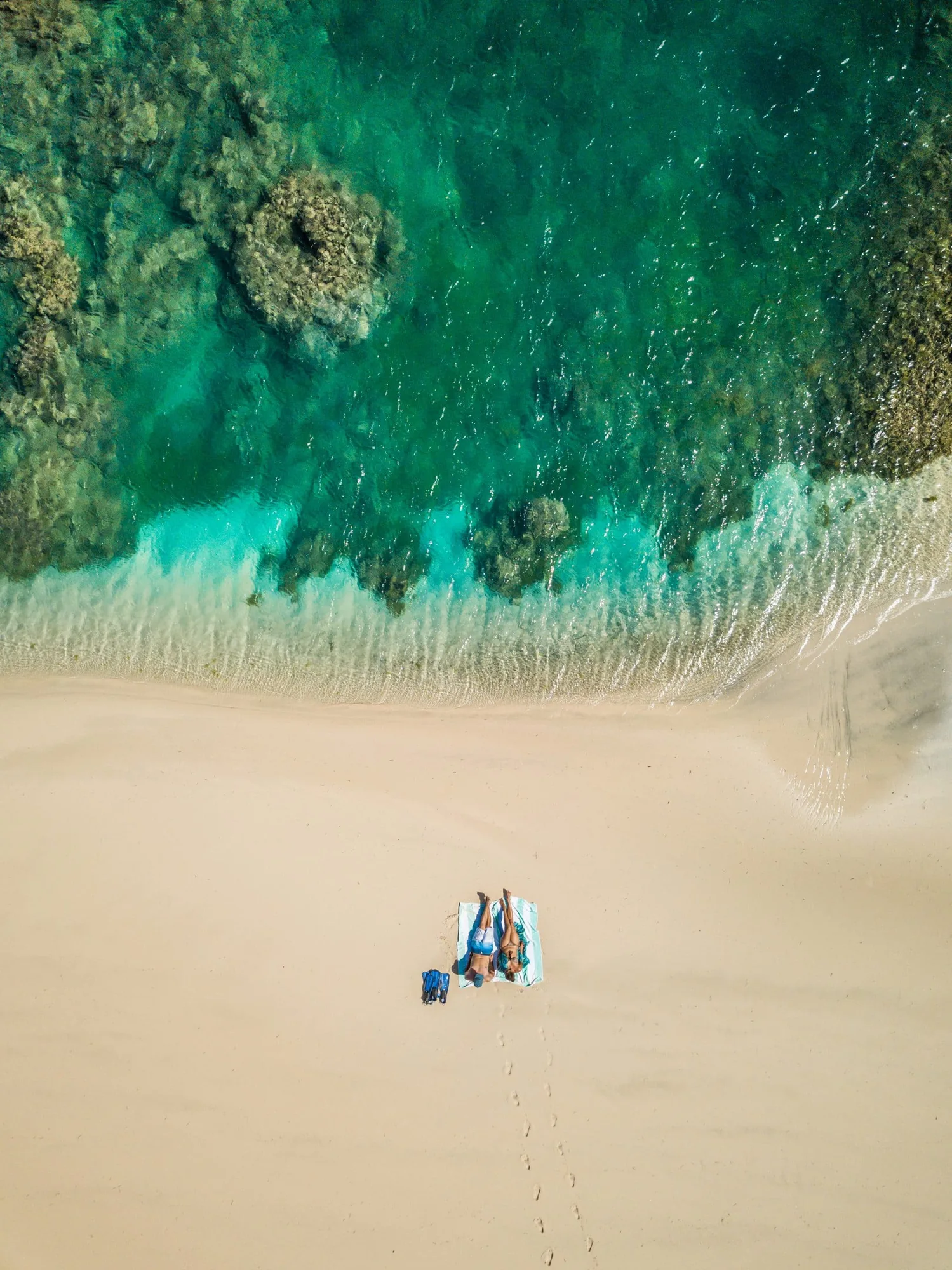 A near-vertical drone view of a white sand spit meeting turquoise water, with two people lying on towels and a line of footprints in the sand.