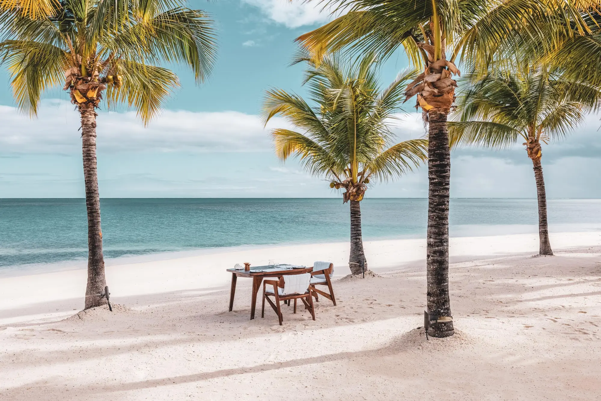 A small timber table laid for two, with two chairs, on white sand between leaning coconut palms, with flat turquoise sea and a low bank of cloud behind.