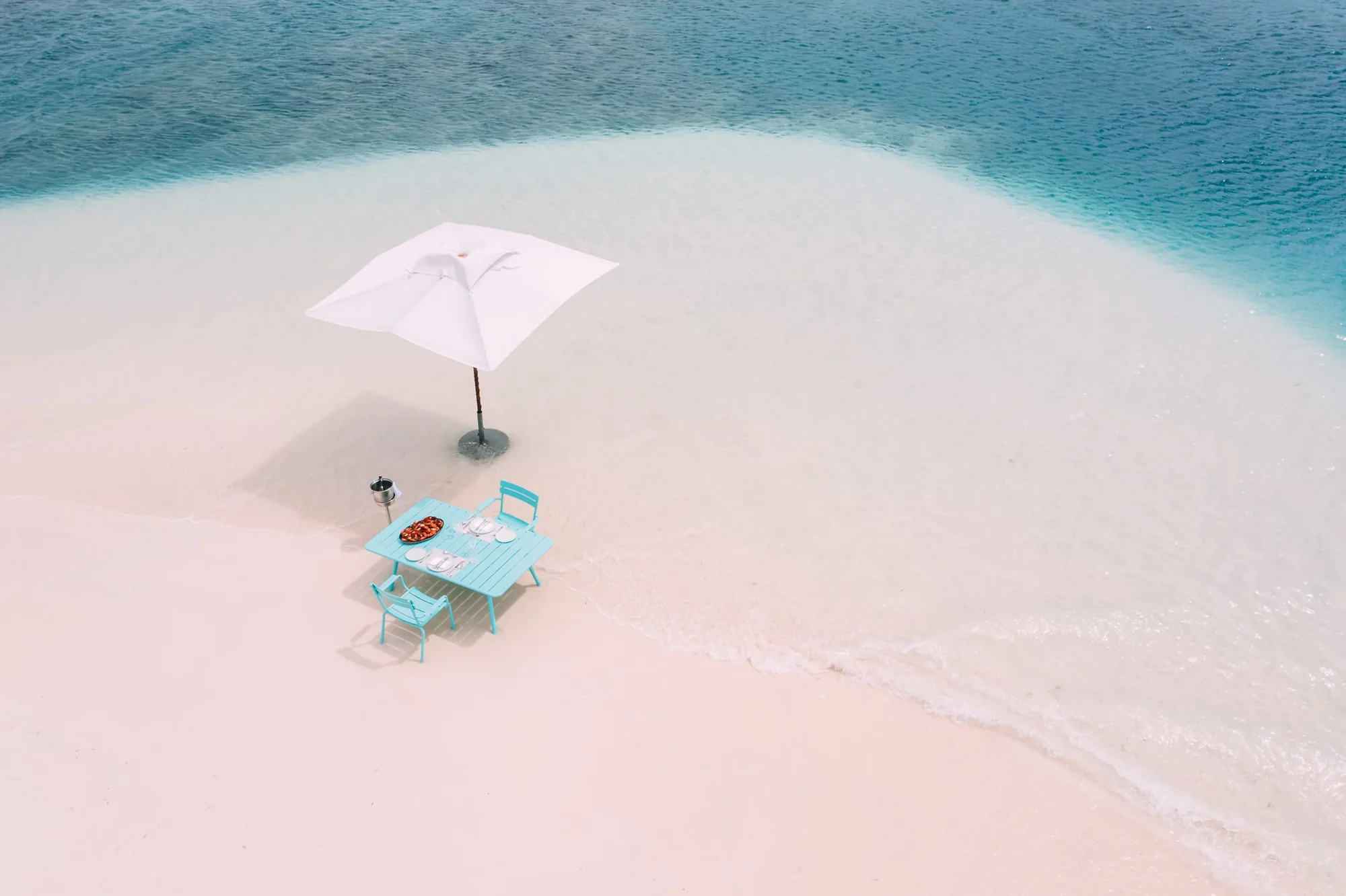 Seen from above and at a distance: a single white parasol and a turquoise table laid for two, alone on an empty pale sandbar ringed by shallow turquoise water.