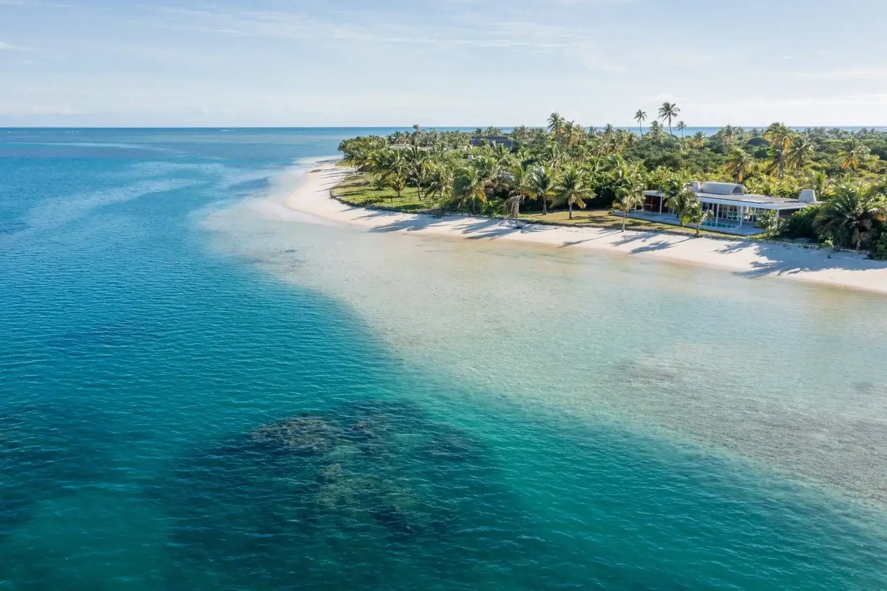 An aerial view across deep blue water to a curving white beach fringed with dense palms, a low pale building among the trees, and a dark reef patch in the turquoise shallows.