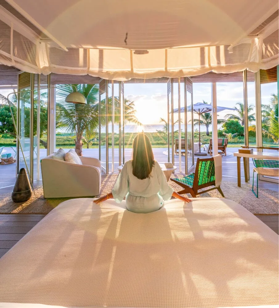 A person in white seated on a bed, seen from behind, under a curved pavilion roof, looking out through open glass to a potted palm, deck furniture and the sea in low sun.