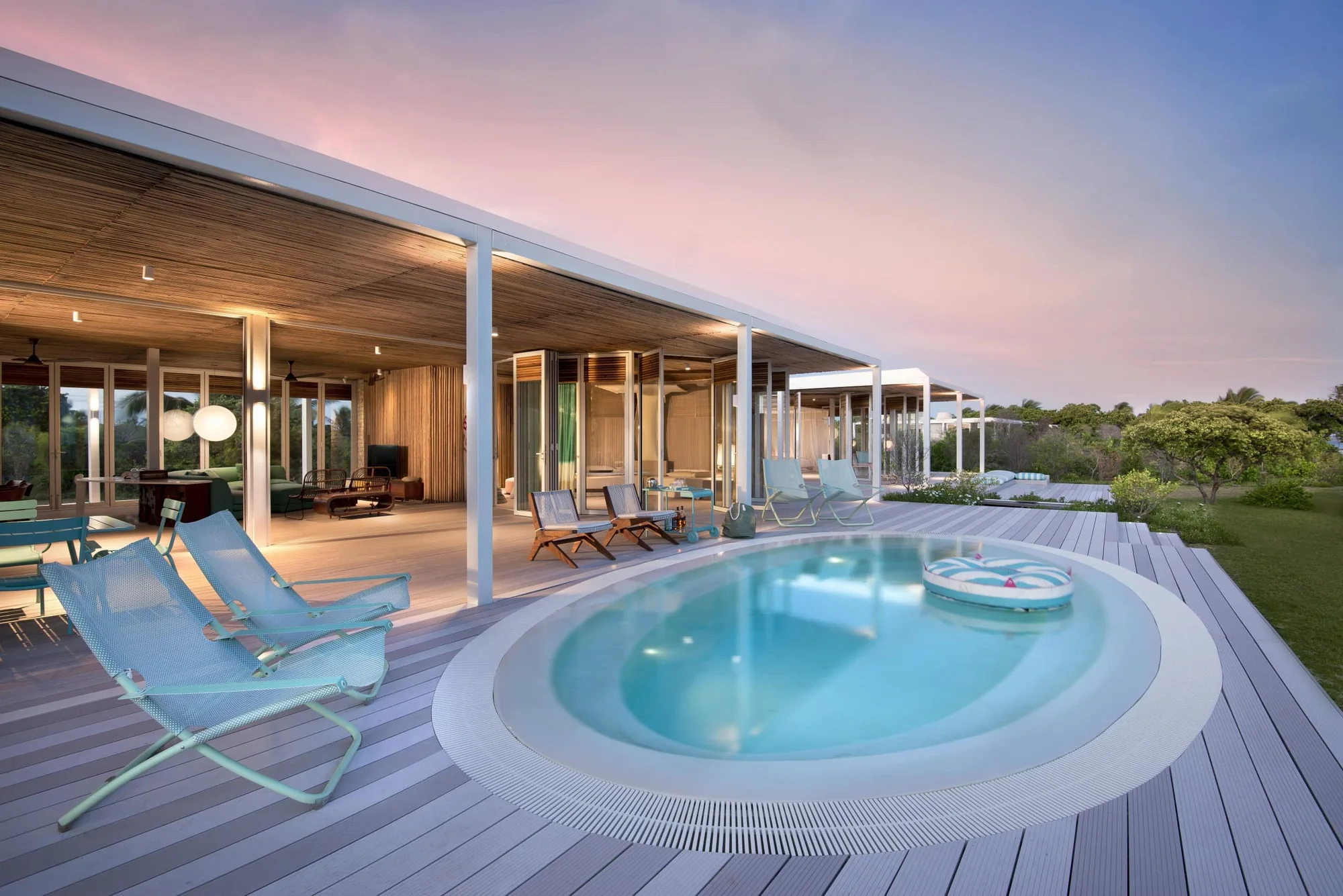 A long low villa at dusk with a lit interior behind glass walls, fronted by a broad deck with a round plunge pool and pale-blue loungers, under a pink and blue sky.