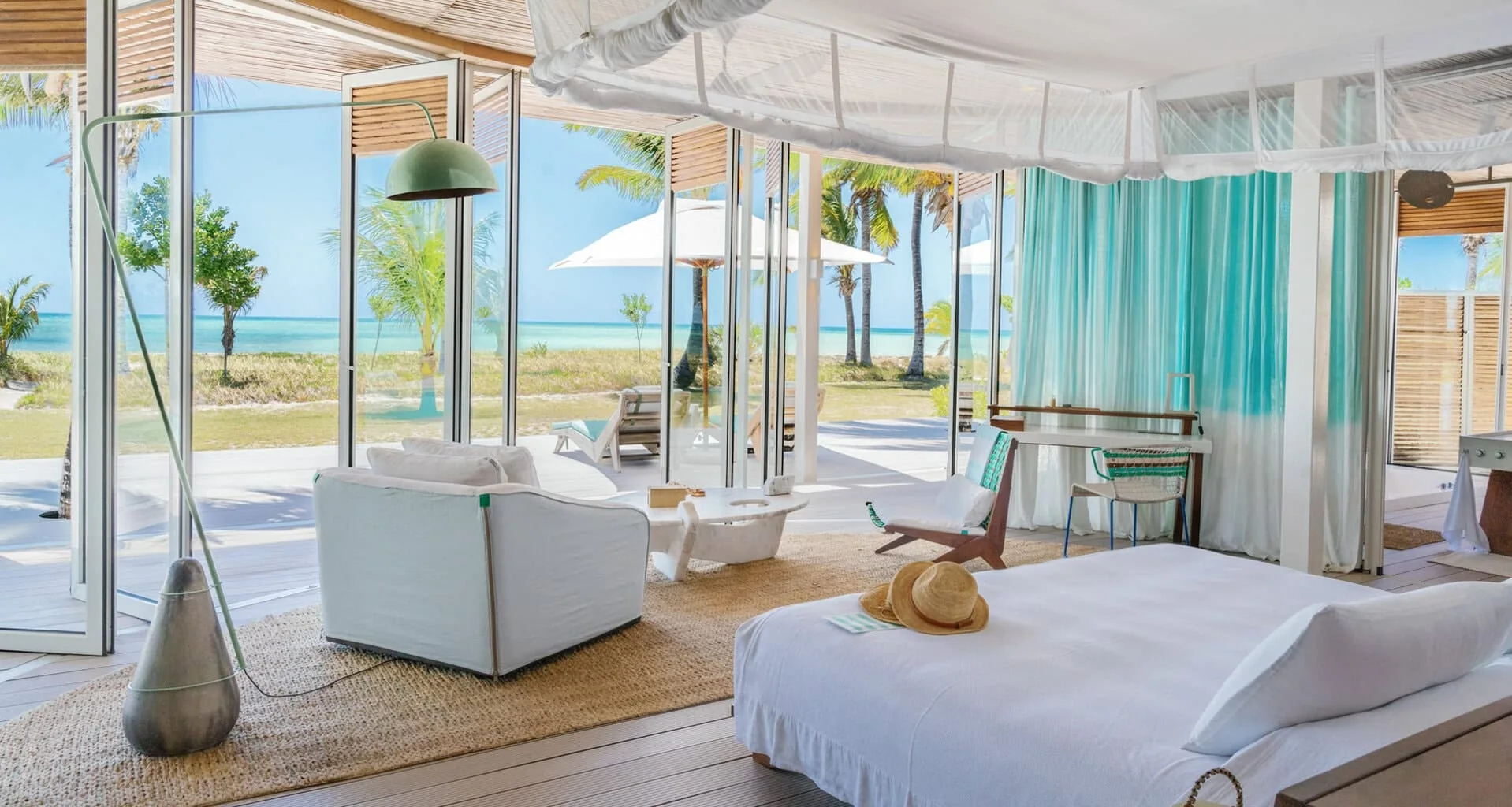 An open-plan villa interior — a bed under a white canopy net, a white armchair and a sisal rug — with folding glass walls opened onto a deck of loungers and parasols, palms and turquoise sea beyond.